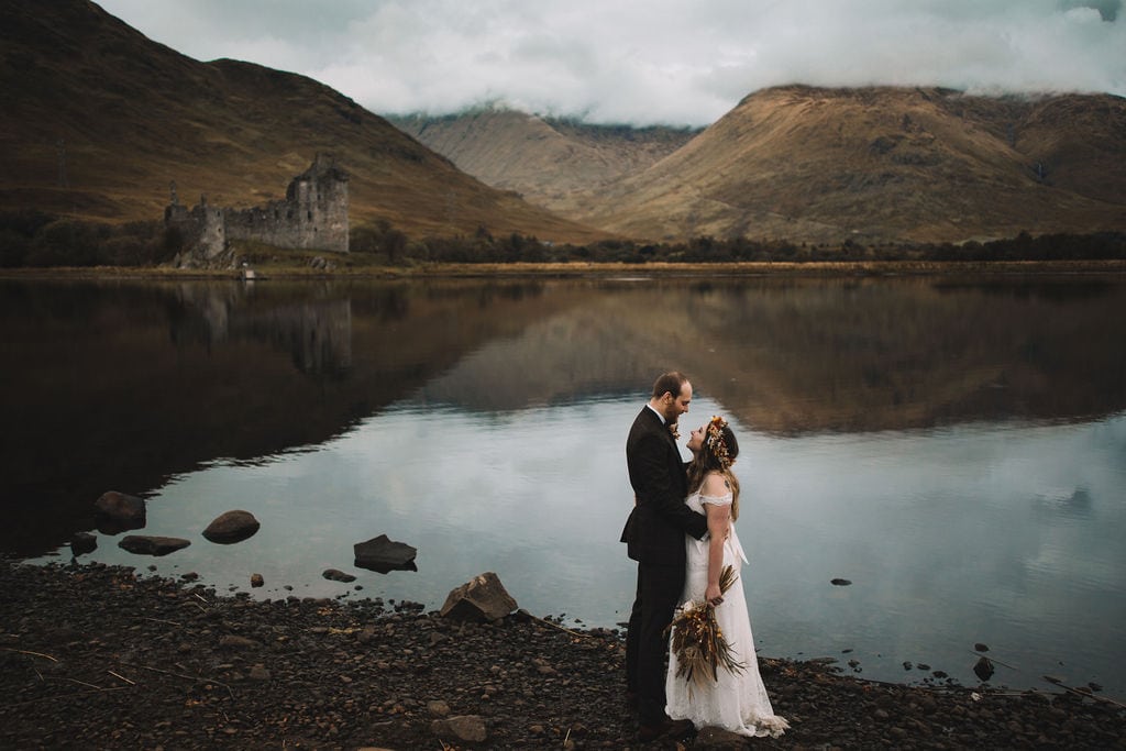 Scottish Highland Wedding, couple cuddling in front of loch awe with a castle in the background. Scotland Elopement, couple cuddling in front of kilchurn castle on loch awe