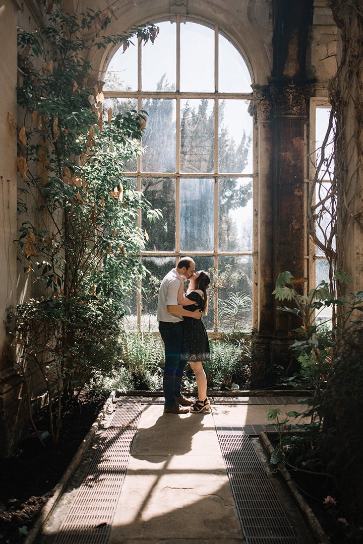 Couple cuddling in front of a window in castle Ashby's orangey with greenery all around them during couple session