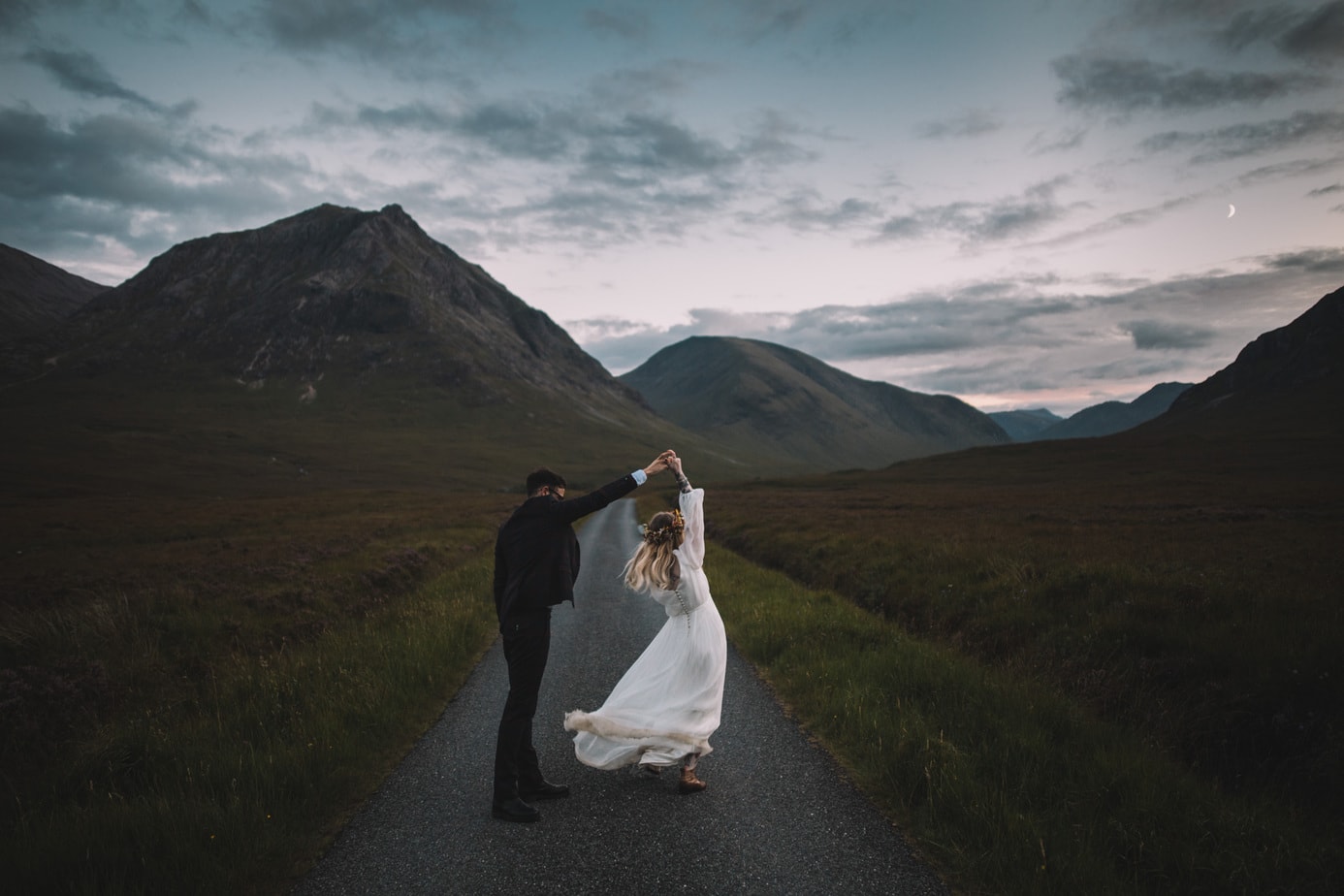 Set on the road to Glen Etive, A bride is twirling around her groom at blue hour and a little moon can be seen between the hills.