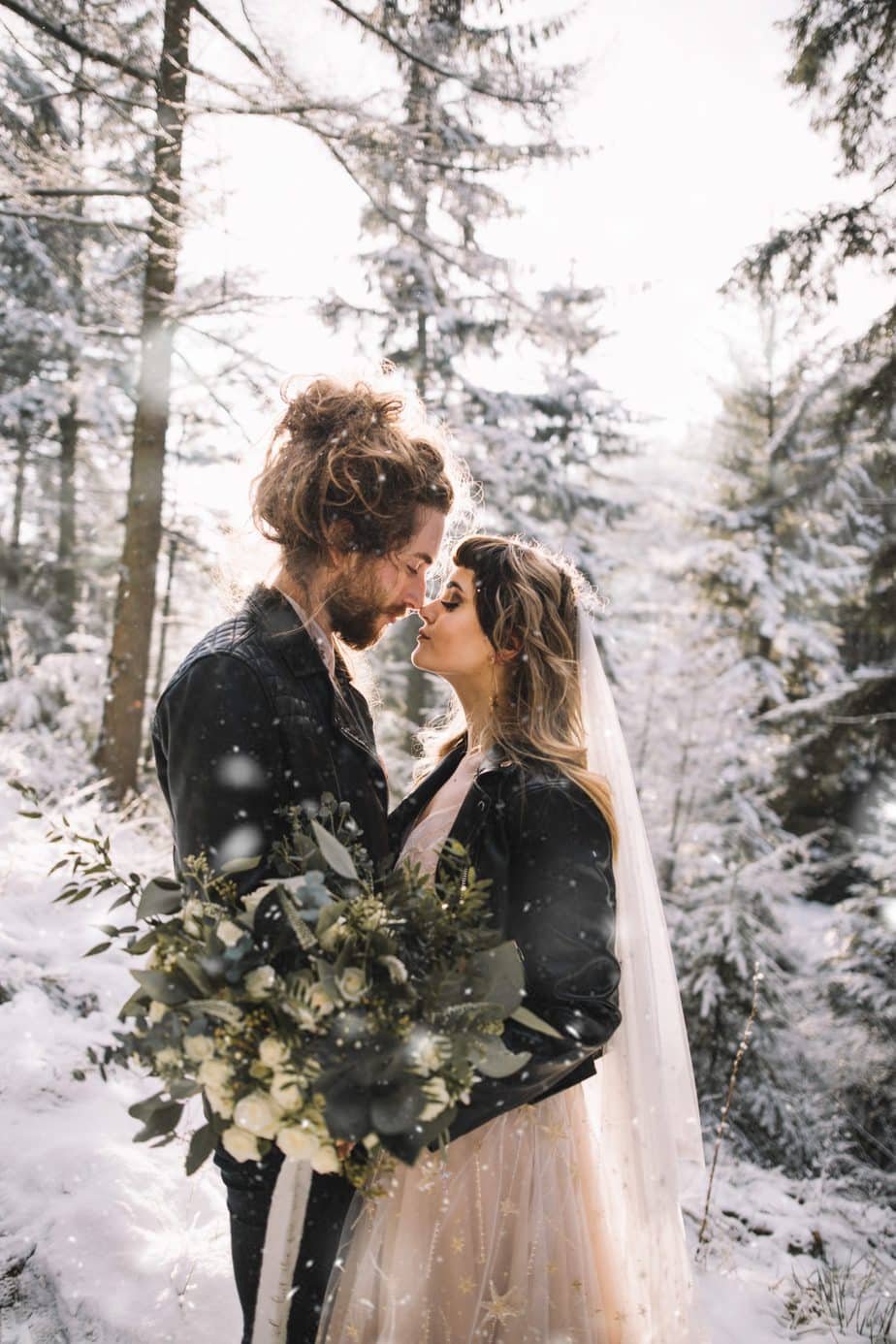 Couple touching noses in a snowy woodland with snow falling around them after they eloped. Bride wearing willowy celestial dress and holding a wild bouquet in green and white tones.