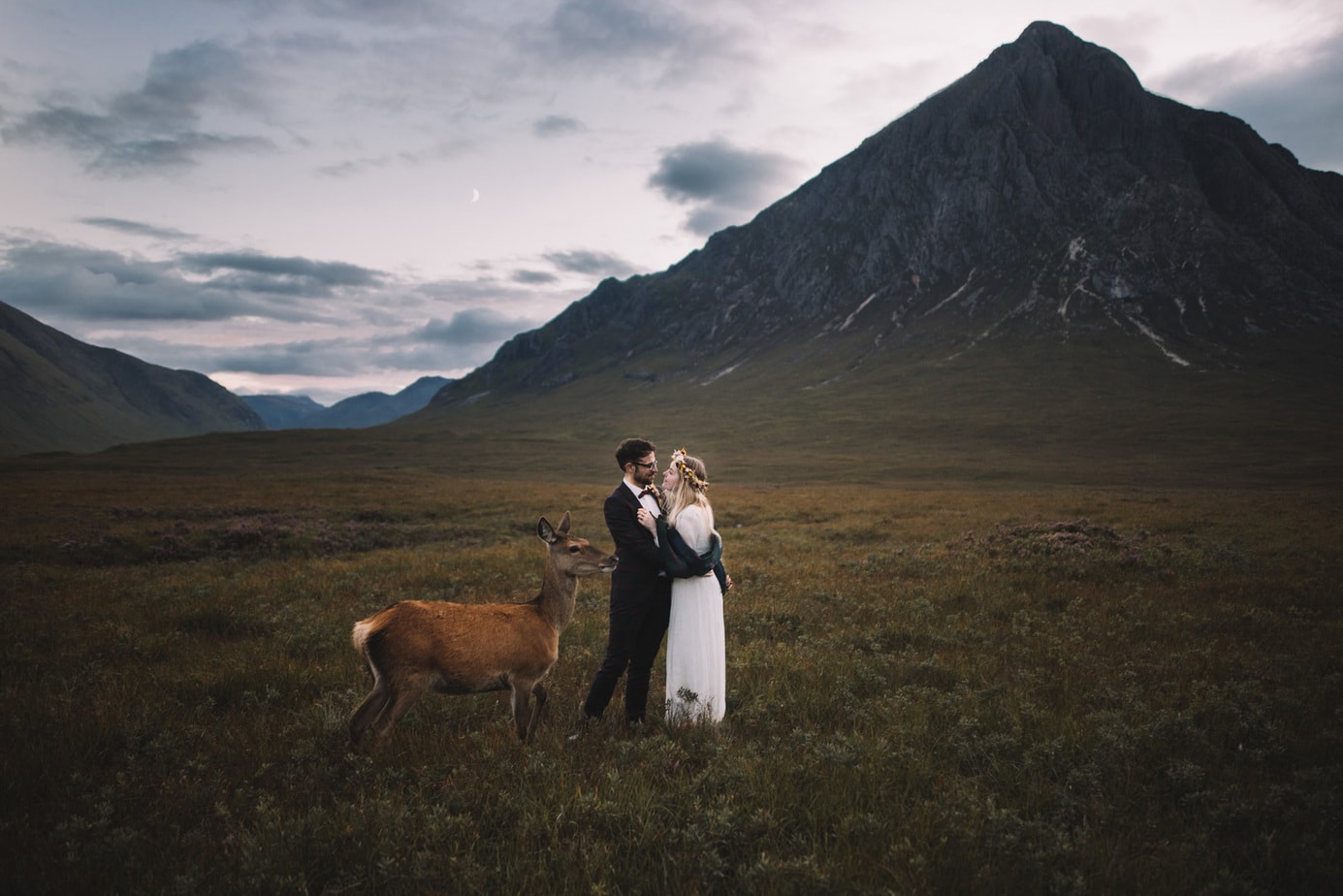 Scotland Elopement. Set in front of Glen Etive, a couple are cuddling during their elopement pictures while a deer is stood with them.