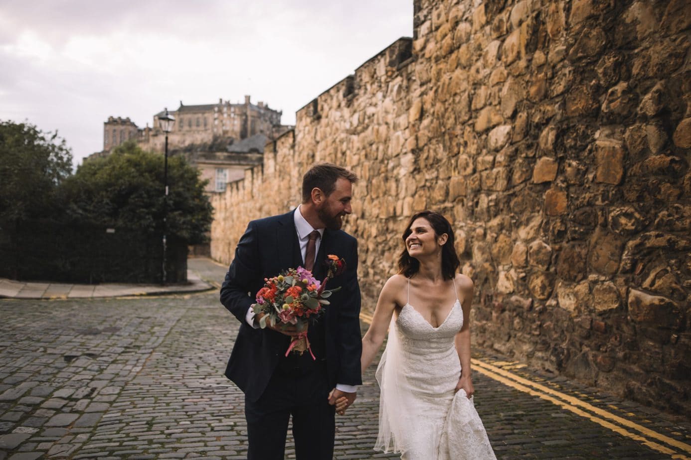 Couple walking up a cobbled street smiling at each other with Edinburgh castle in the background during their elopement
