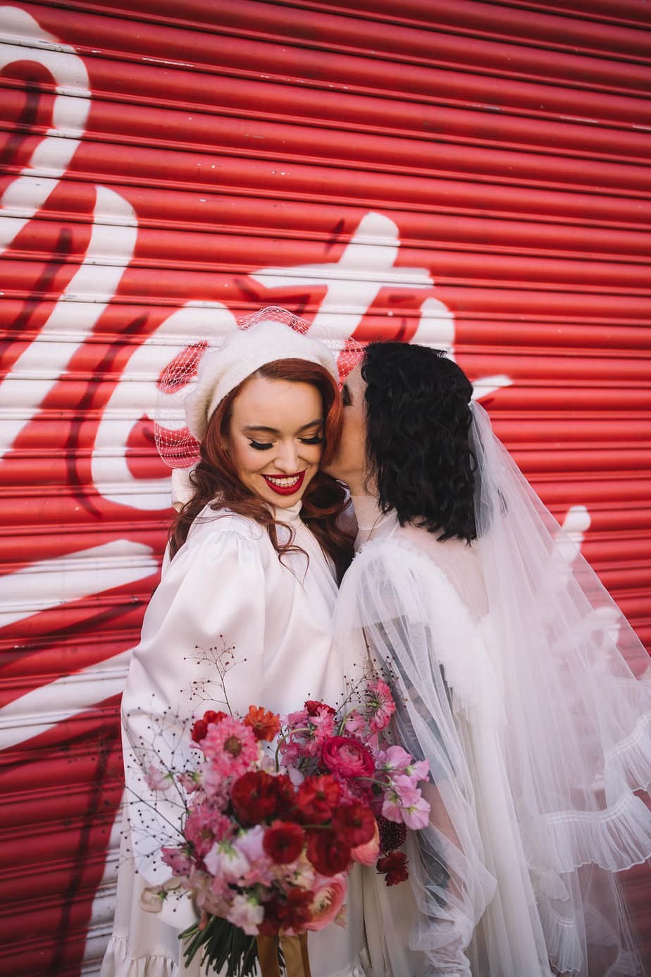 Manchester City centre elopement with two brides. Bride Esme is being kissed by Becca in front of a red graffiti shutter