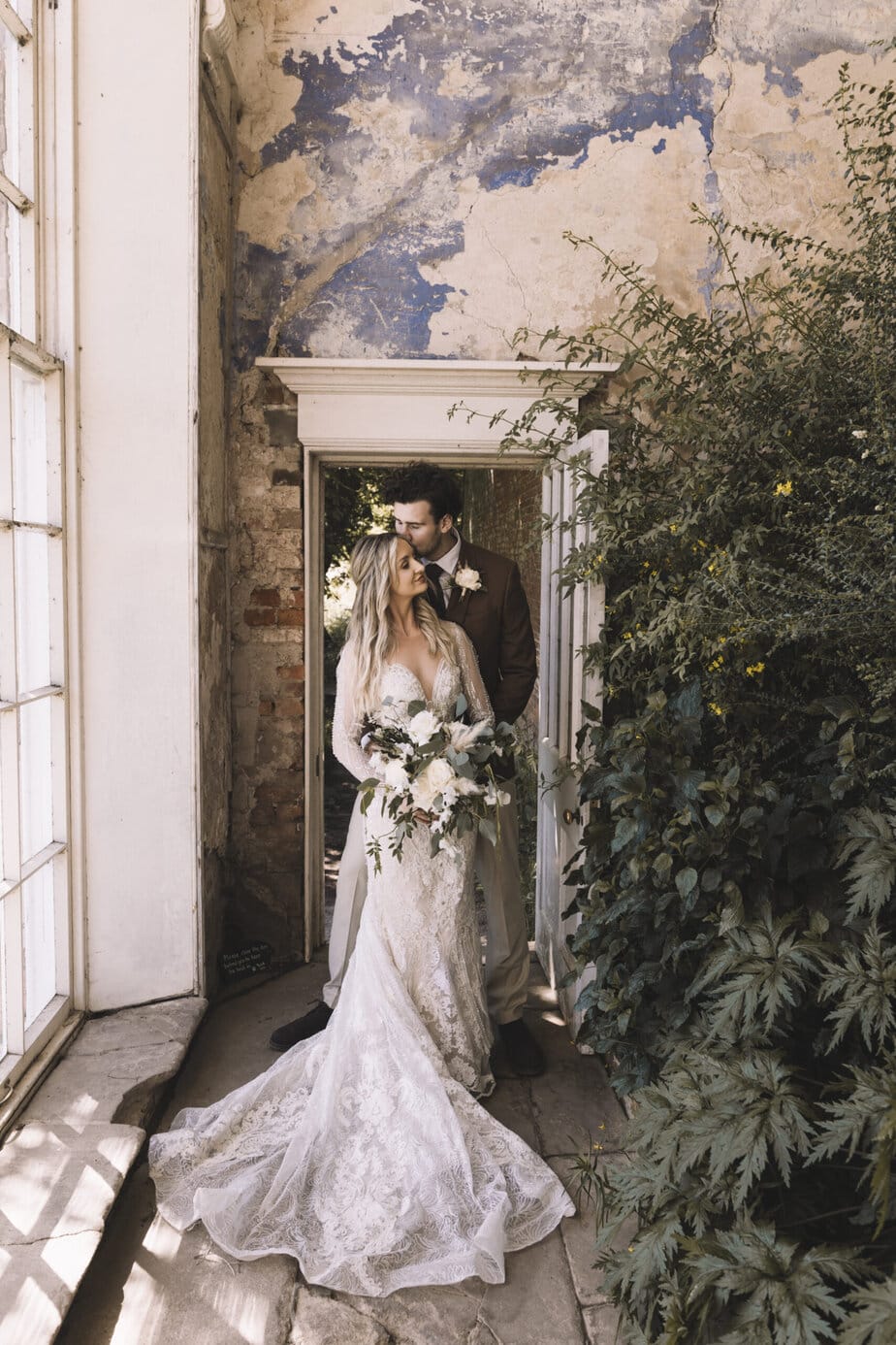 Groom kissing bride on the cheek as they stand in the doorway of Calke Abbey's orangery on their wedding day.