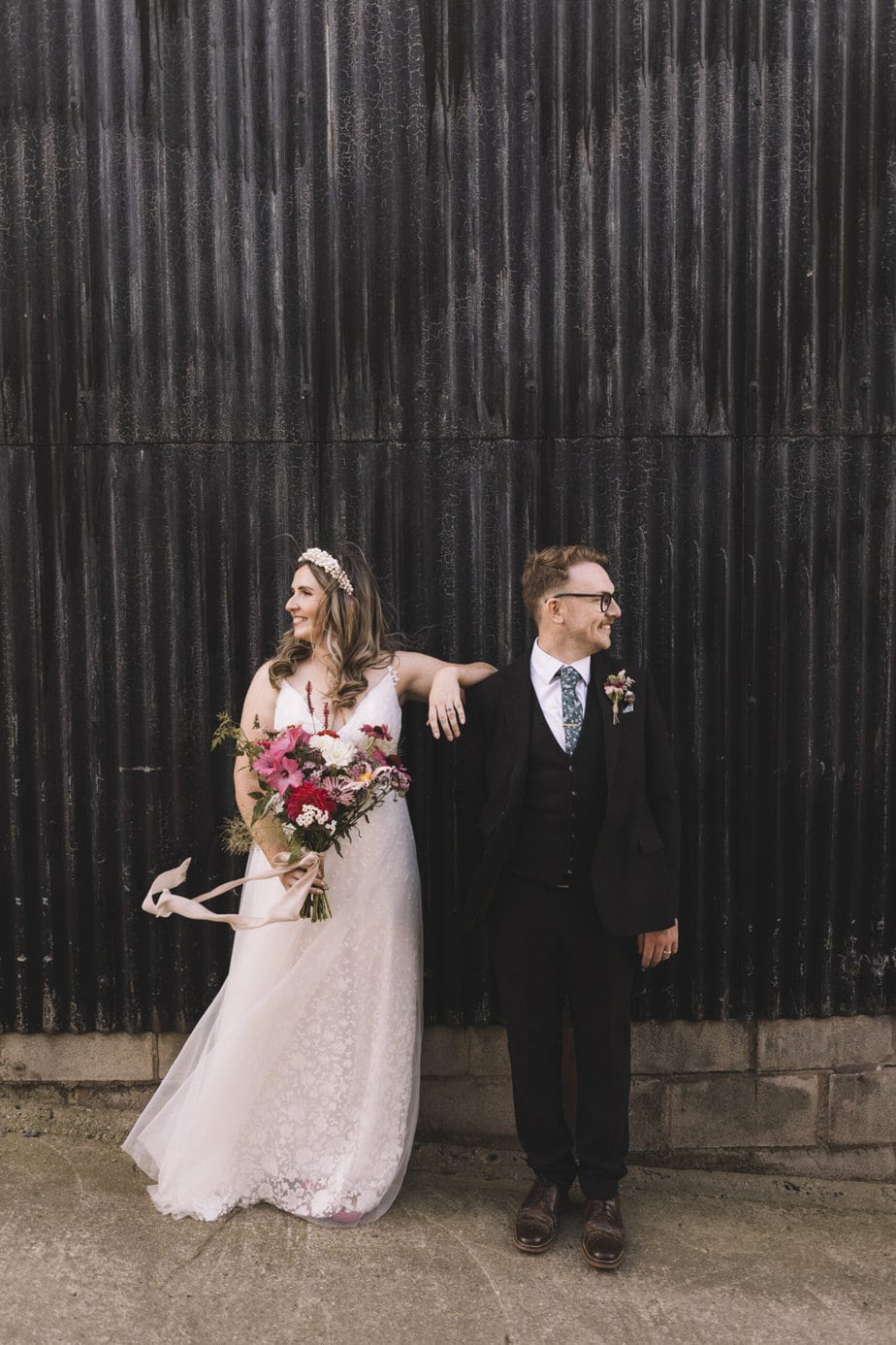 Bride leans on groom, both looking in opposite directions with the black barn wall behind at their grange farm wedding